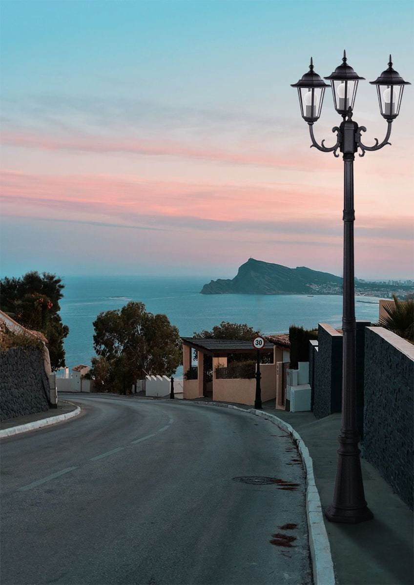 Czarna lampa ogrodowa stojąca Standard H233 z trzema oprawami na żarówki E27. Produkt ma klasyczny, metalowy kształt z ozdobnymi detalami, ustawiony na prostym, prostokątnym cokole. Widoczna w otoczeniu nadmorskim, na zakręcie drogi.