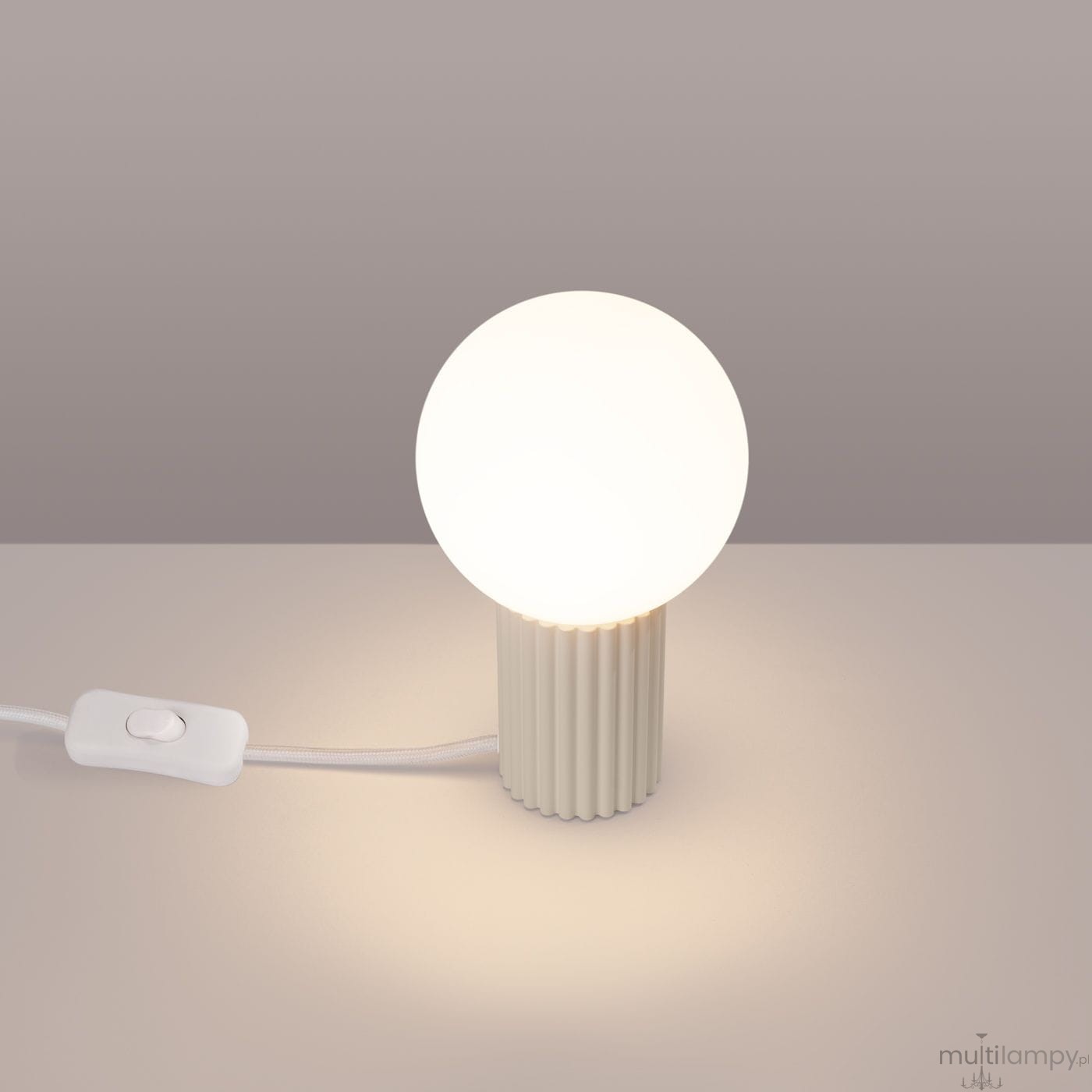 Beżowa lampa stołowa Halo z okrągłym, białym kloszem, umieszczona na cylindrycznej, żebrowanej podstawie. Widoczny kabel z włącznikiem. Produkt ujęty frontalnie na jasnym tle.