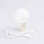 Beżowa lampa stołowa Halo z okrągłą, białą kloszem i żebrowanym stojakiem. Widoczny biały przewód z włącznikiem. Lampa z gniazdem na żarówkę G9, ustawiona na białym tle.