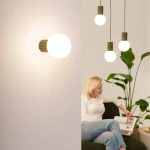 Kinkiet Halo 1xG9 IP44 w kolorze oliwkowym, z okrągłą, białą kloszem. Ujęcie frontalne, zamontowany na ścianie. W tle widoczna osoba siedząca na kanapie, otoczona roślinami, co podkreśla domowy kontekst.