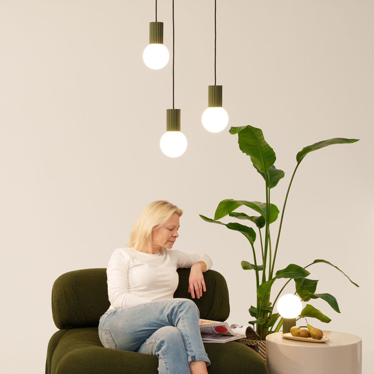 Lampa wisząca Halo 1xG9 w kolorze oliwkowej zieleni z trzema okrągłymi źródłami światła. Zawieszona na czarnych linkach, umieszczona nad osobą siedzącą na zielonej kanapie, w otoczeniu rośliny doniczkowej. Tło jasne, minimalne.