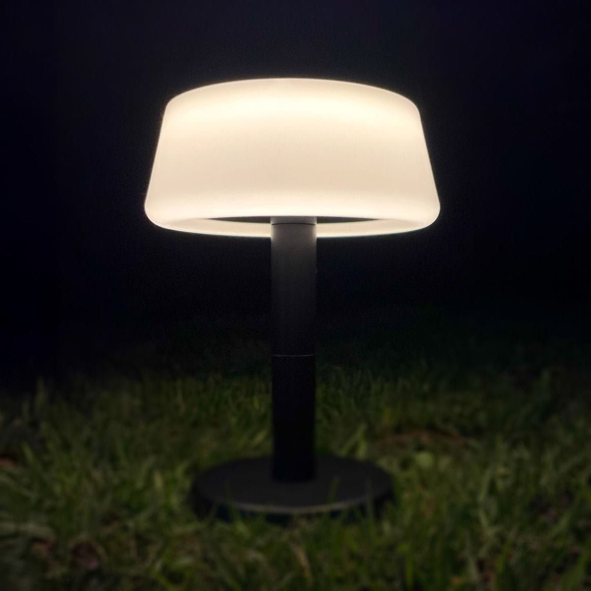 Lampa stołowa solarna LED Aure, czarna, z okrągłą podstawą i osłoną z matowego tworzywa. Źródło światła 2,2W, temperatura barwowa 2700K, odporność IP44. Ujęcie z boku, lampa z włączonym światłem, umieszczona na trawie w ciemnym otoczeniu.