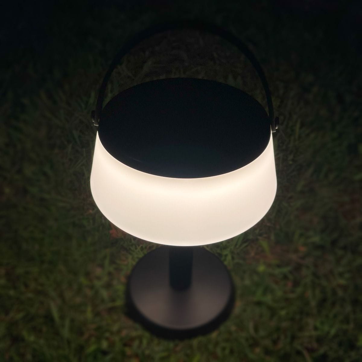 Czarna lampa stołowa solarna LED Aure o mocy 2,2W, z białym kloszem w kształcie półkuli. Ujęcie z góry, stojąca na trawie. Oświetlenie o temperaturze barwowej 2700K, klasyfikacja IP44, odporna na warunki atmosferyczne.