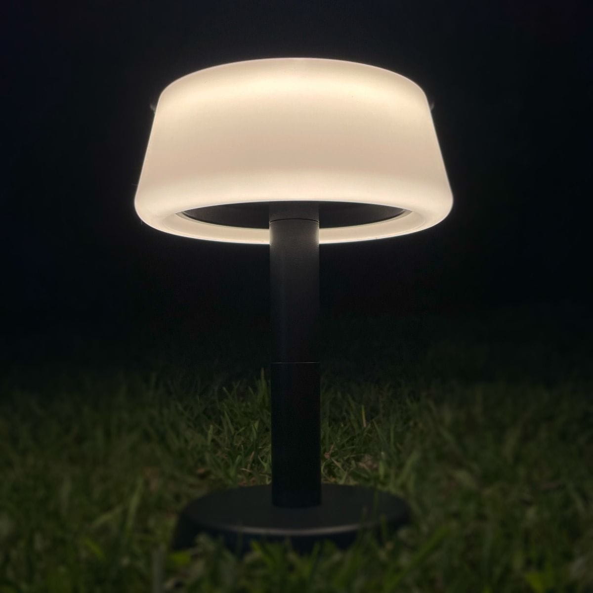 Lampa stołowa solarna LED Aure o mocy 2,2W, kolor czarny. Posiada okrągłą, białą klosz, umieszczoną na prostym słupku. Widok z dołu, na tle trawy, podkreślający subtelne oświetlenie. Klasa szczelności IP44 zapewnia odporność na warunki atmosferyczne.