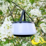 Lampa stołowa solarna LED Aure o mocy 2,2W, kolor czarny, z białym kloszem. Zawieszona na gałęzi wśród białych kwiatów, z widocznym czarnym uchwytem. Oświetlenie w ciepłej barwie 2700K, stopień ochrony IP44.