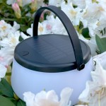 Lampa stołowa solarna LED Aure o mocy 2,2W. Posiada czarną górną część z panelem słonecznym i uchwyt. Korpus jest biały, matowy. Umieszczona pośród białych kwiatów, co podkreśla jej zewnętrzny charakter. Wodoodporność IP44.