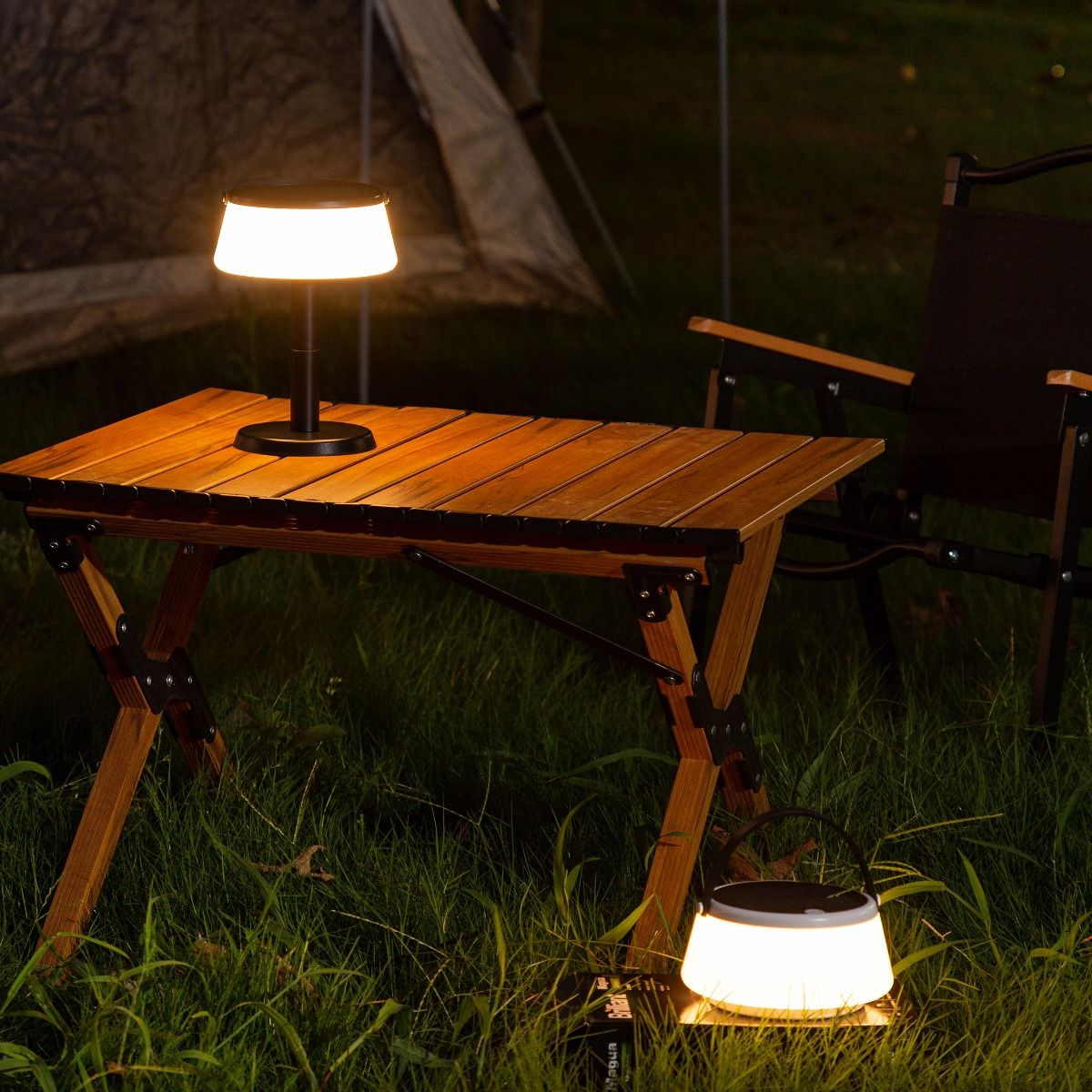 Czarna lampa stołowa solarna LED Aure o mocy 2,2W i temperaturze barwowej 2700K, ustawiona na drewnianym stole w ogrodzie. W tle widoczny składany fotel i namiot, otoczenie zielone, z trawą. Lampa emituje ciepłe światło, tworząc przytulną atmosferę.