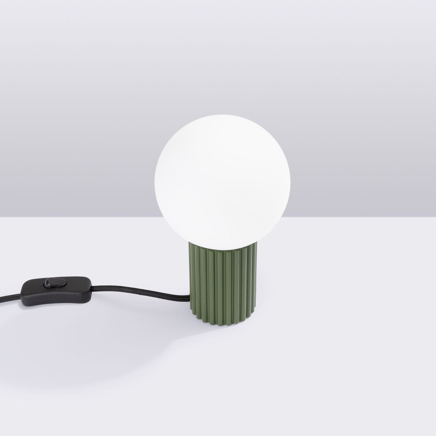 Lampa stołowa Halo w kolorze oliwkowej zieleni z okrągłą, białą kloszem. Posiada teksturowaną podstawę w pionowe żłobienia oraz czarny przewód z włącznikiem. Ujęcie z boku, na jasnym tle.