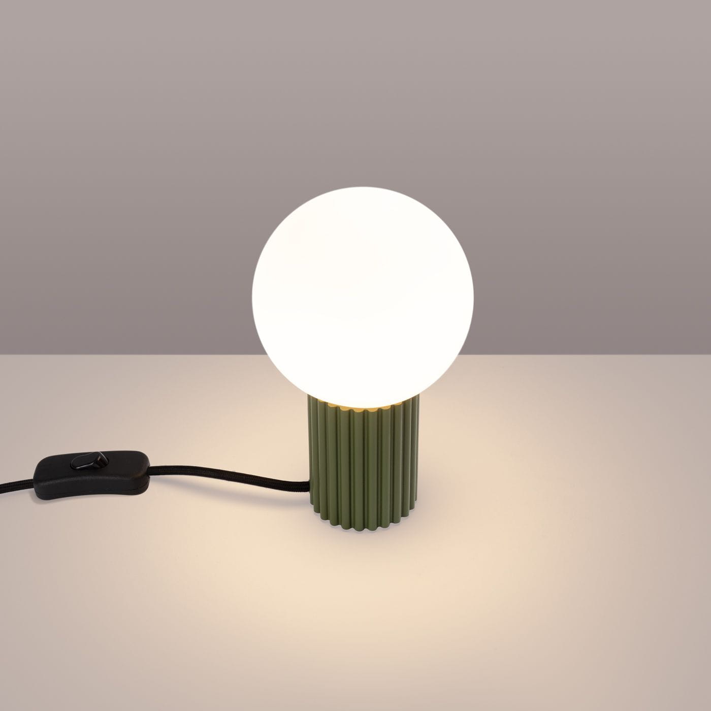 Lampa stołowa Halo w kolorze zielonej oliwki, z okrągłą, białą żarówką G9. Ustawiona na płaskiej powierzchni, z widocznym czarnym kablem i włącznikiem. Podstawa o fakturze żłobkowanej, tworzy minimalistyczny i nowoczesny design.