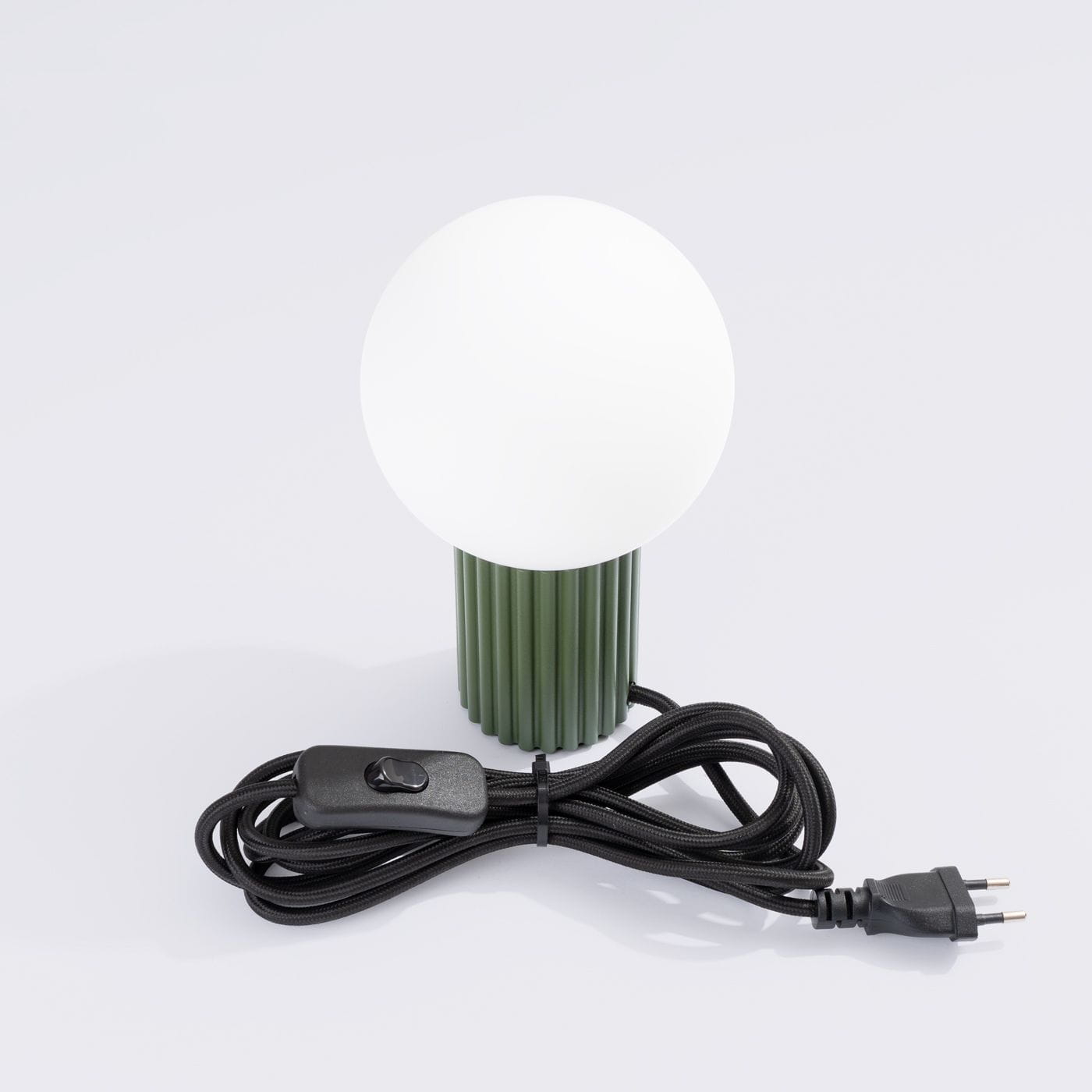 Lampa stołowa Halo 1xG9 w kolorze oliwkowej zieleni, z białą, okrągłą kloszem. Ustawiona w pionie, z widocznym czarnym kablem z przełącznikiem. Dół lampy ma teksturowaną, żebrowaną strukturę.