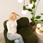 Lampa stołowa Halo w kolorze oliwkowym, z podstawką w teksturze pionowych żłobień. Oświetlenie z kulistą żarówką G9, umieszczona obok rośliny i nowoczesnego stolika. Ujęcie z boku, z osobą siedzącą na zielonej kanapie.