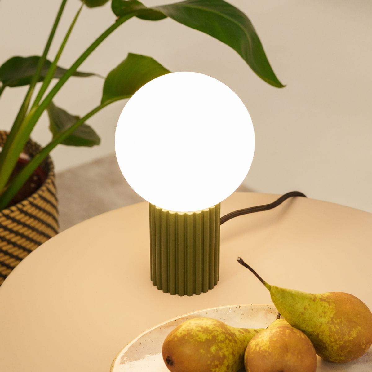 Lampa stołowa Halo w kolorze oliwkowej zieleni z okrągłą, białą żarówką na cylindrycznej podstawie. Produkt stoi na jasnym stole, obok widoczne gruszki. Kabel zasilający wychodzi z boku lampy.