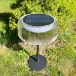 Lampa stołowa solarna LED 2,5W 3000K IP44 w kolorze czarnym, z przezroczystym kloszem w żłobienia. Ustawiona na metalowej podstawie, z panelem słonecznym na górze. W otoczeniu zieleni, na trawie.