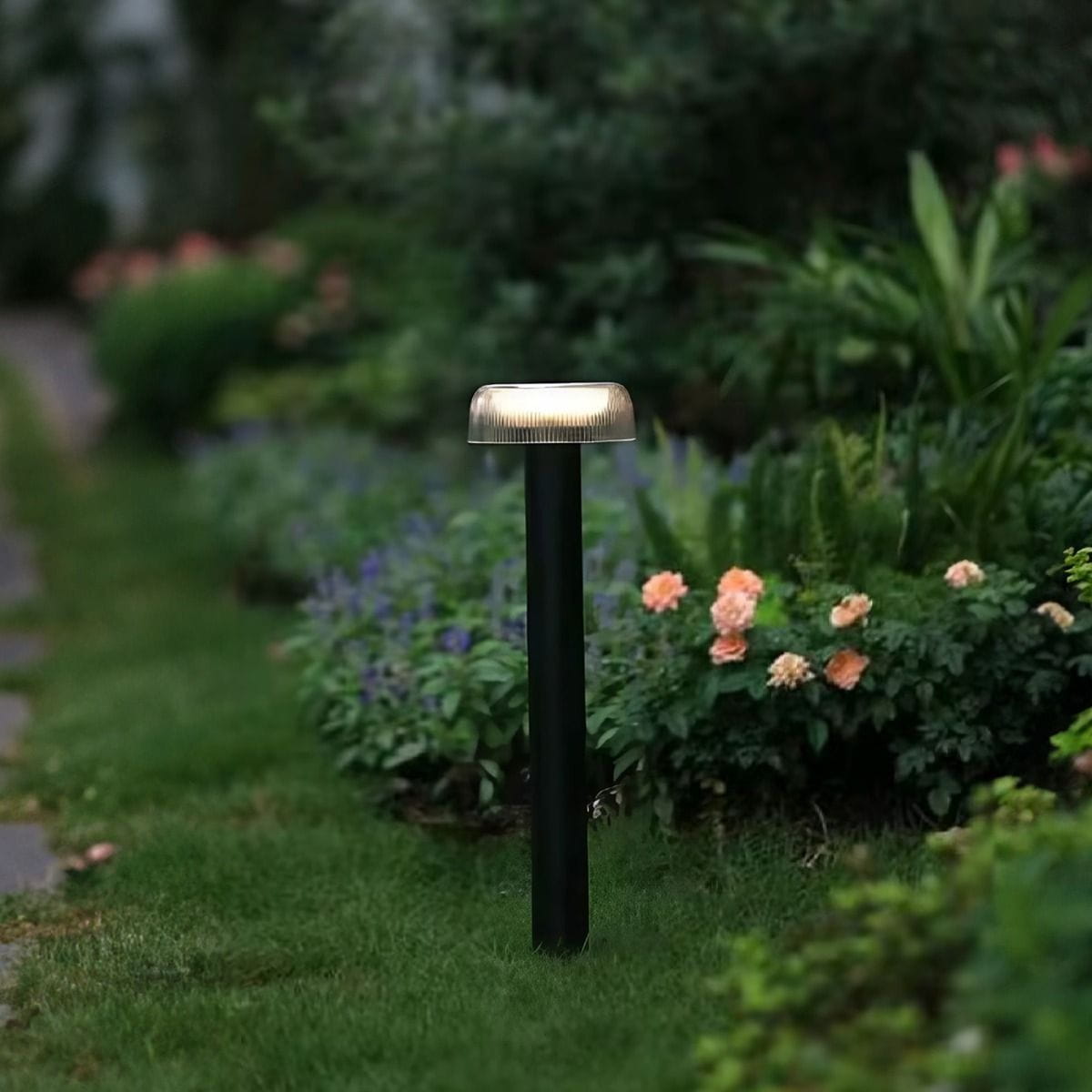 Lampa ogrodowa solarna Bluet, LED 2,5W, 3000K, IP44 w kolorze czarnym. Stoi na trawniku, z przezroczystym kloszem u góry, otoczona zielenią i kwiatami. Produkt w zewnętrznym otoczeniu, w naturalnym świetle.