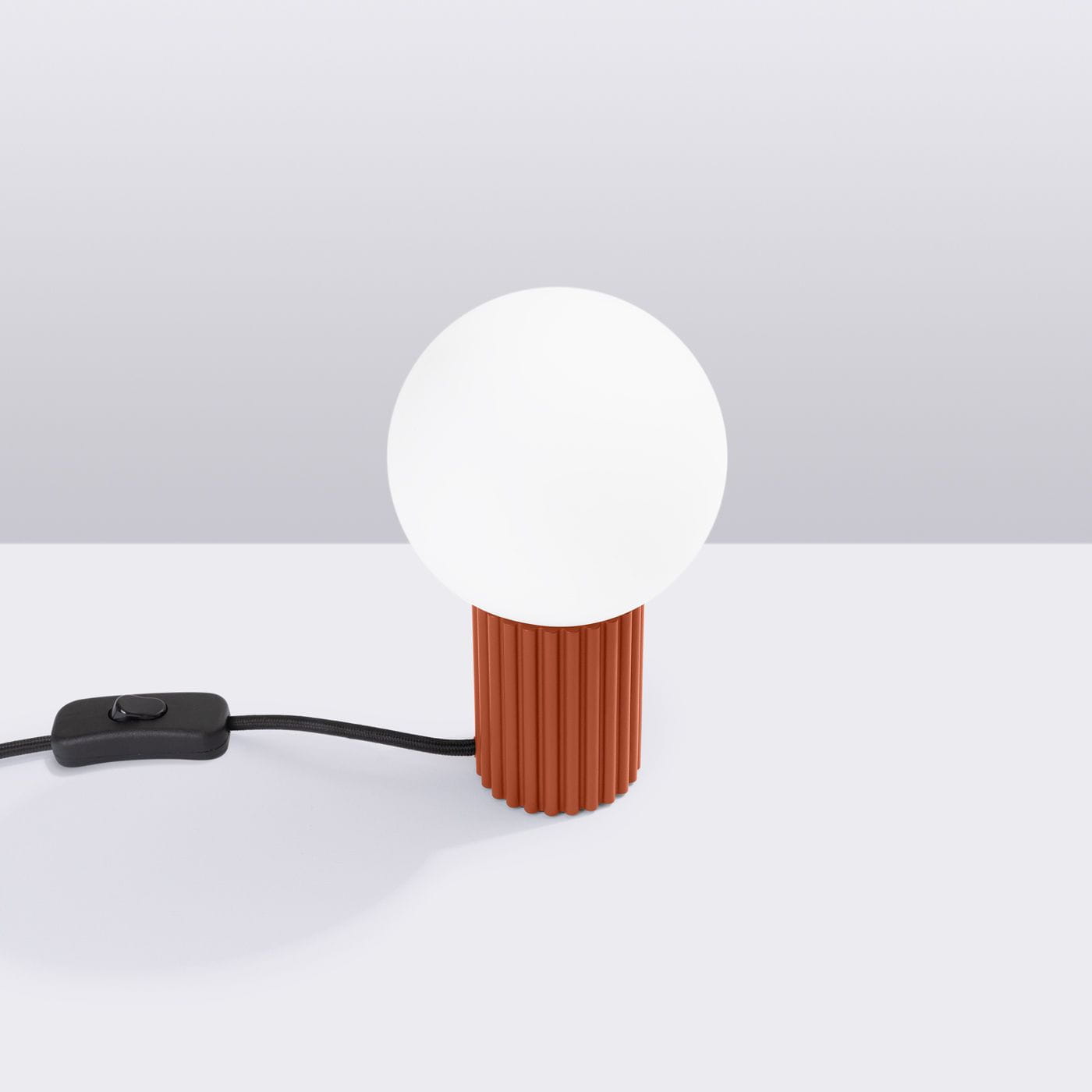 Lampa stołowa Halo w kolorze ochry czerwonej z okrągłą, białą kloszem. Produkt stoi na stole, posiada teksturowaną podstawę z wgłębieniami i czarny kabel z włącznikiem. Tło jasnoszare, proste.