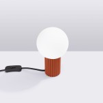 Lampa stołowa Halo w kolorze ochry czerwonej z okrągłą, białą kloszem. Produkt stoi na stole, posiada teksturowaną podstawę z wgłębieniami i czarny kabel z włącznikiem. Tło jasnoszare, proste.