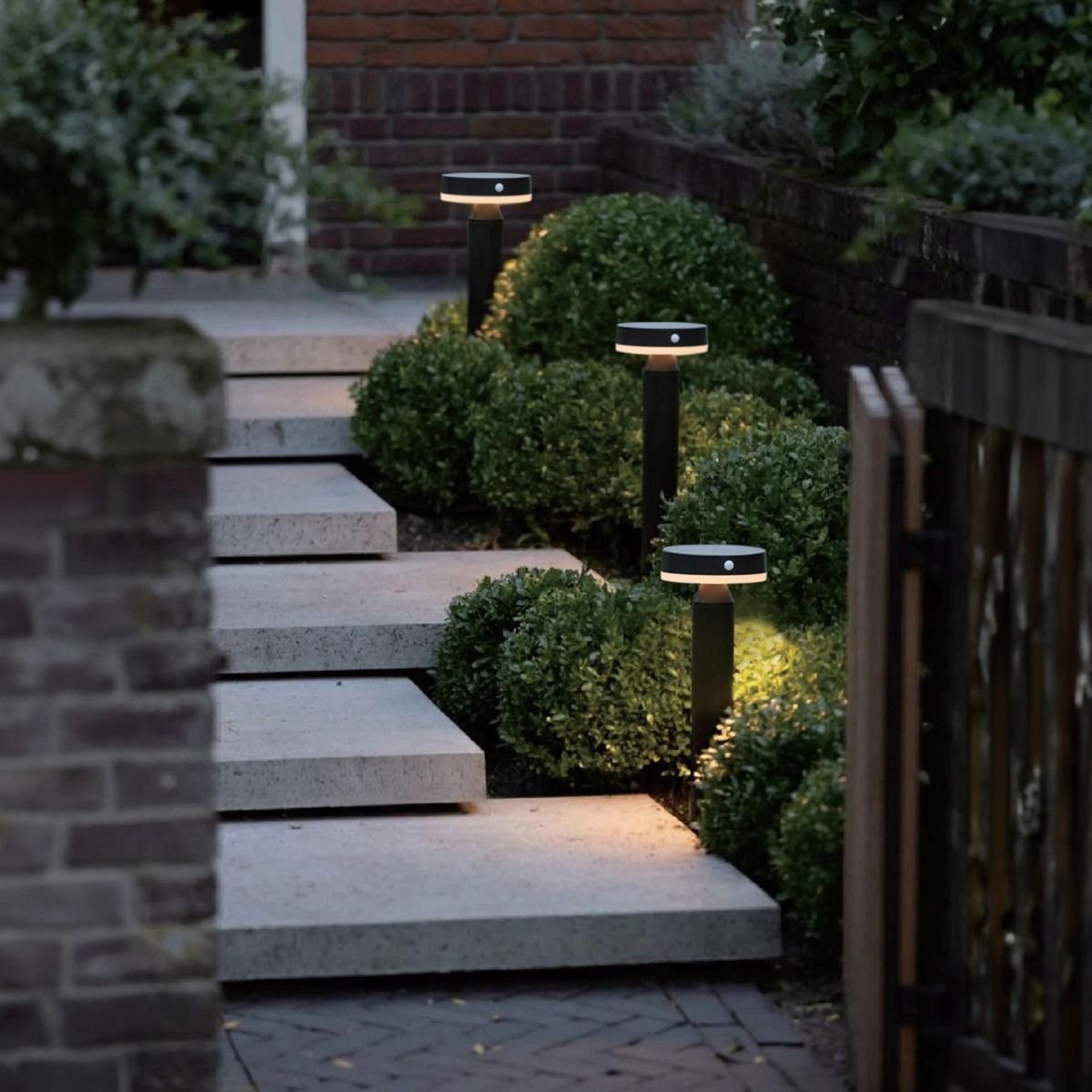 Lampa ogrodowa solarna Carli, LED 4,5W, 3000K, IP44, czarna, umieszczona wzdłuż schodów, o cylindrycznym kształcie z okrągłym kloszem, oświetlająca otoczenie. Tło stanowią krzewy i ceglana ściana domu.