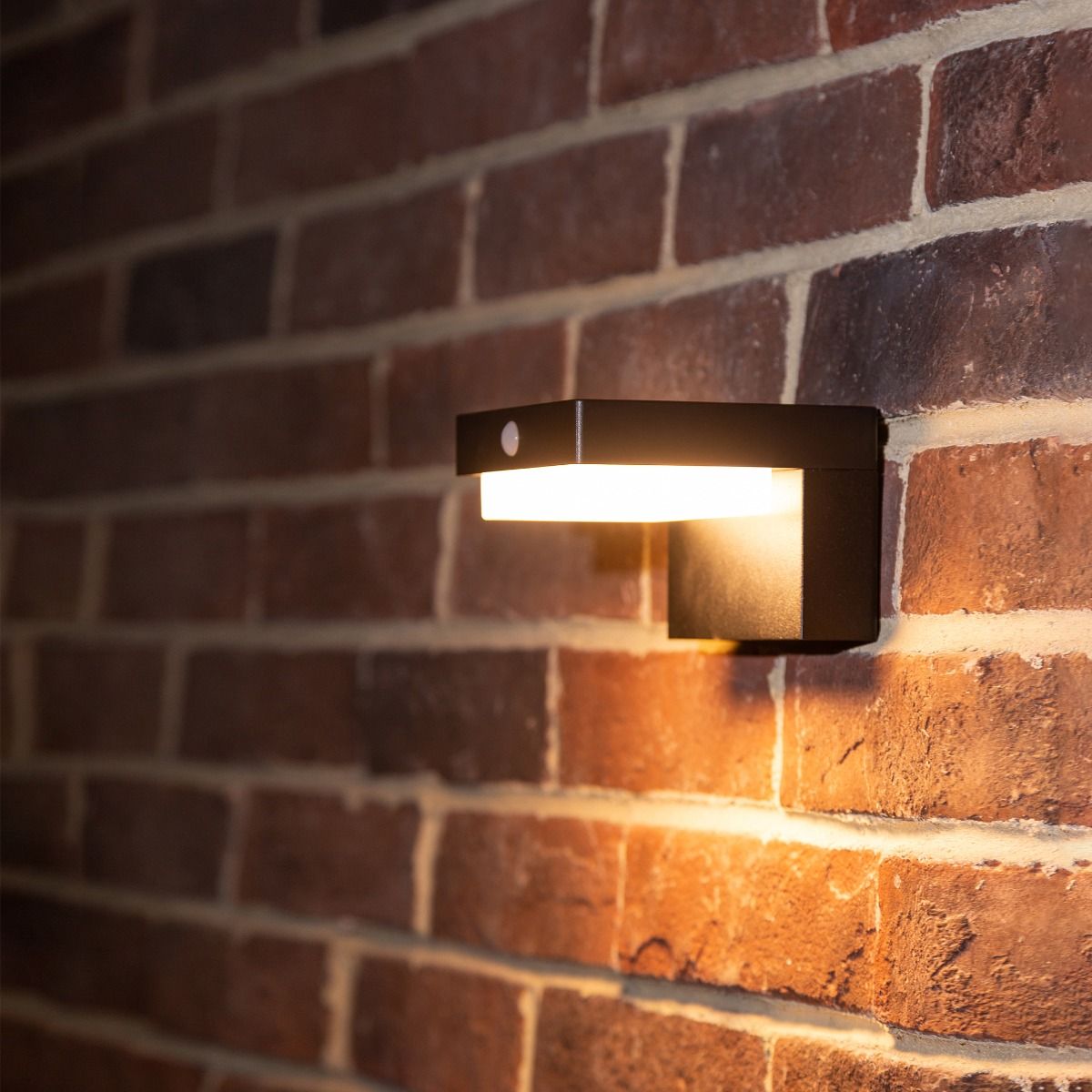 Kinkiet solarny Grid z czujnikiem ruchu LED, moc 4,5W, barwa światła 3000K, IP44, w kolorze czarnym, zamontowany na ścianie z cegły. Oświetlenie skierowane w dół, z widoczną obudową i małym czujnikiem.