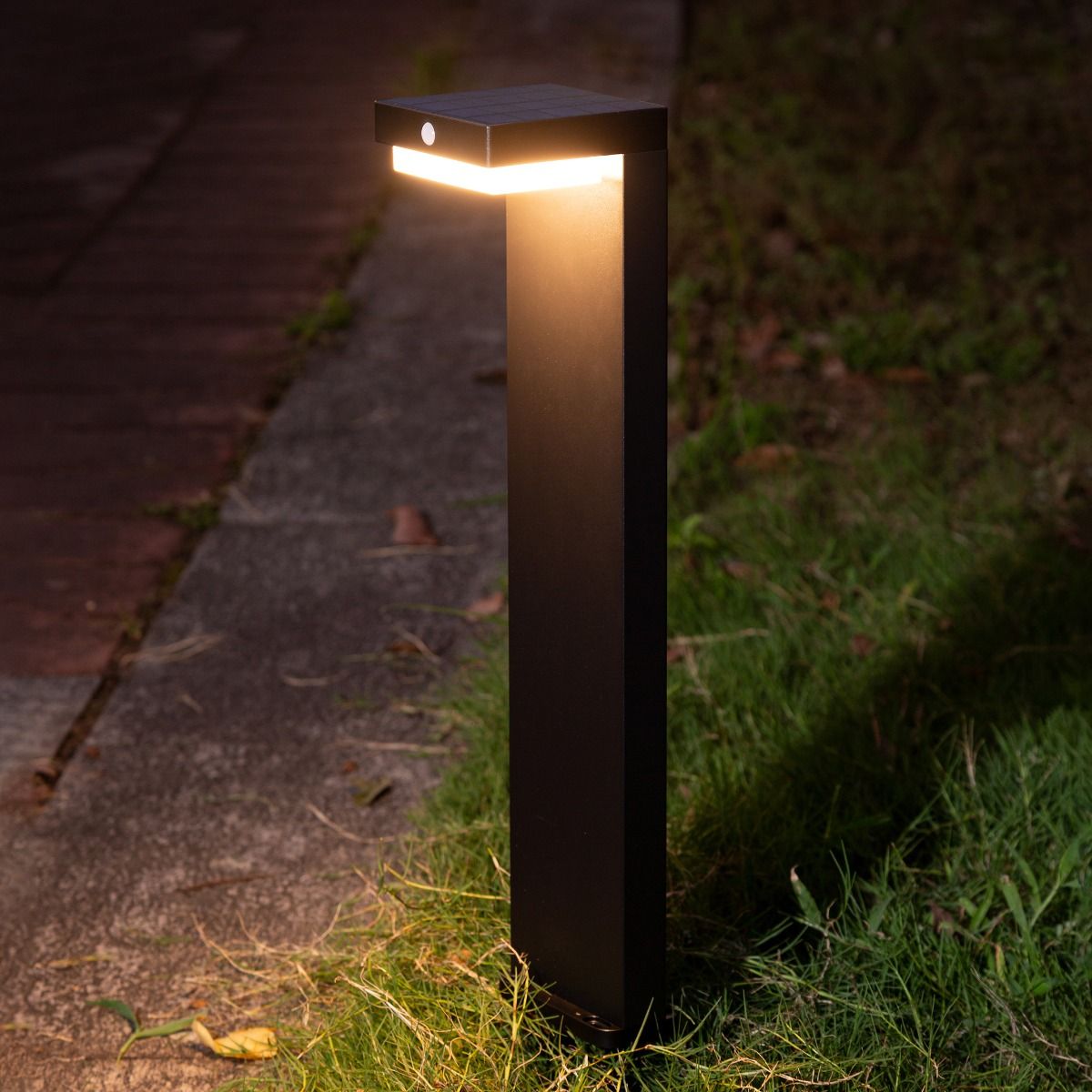 Lampa ogrodowa solarna Grid, czarna, z czujnikiem ruchu, LED 4,5W, 3000K, IP44. Stojąca konstrukcja z prostokątnym kloszem, emitująca ciepłe światło, umieszczona na trawie obok ścieżki.