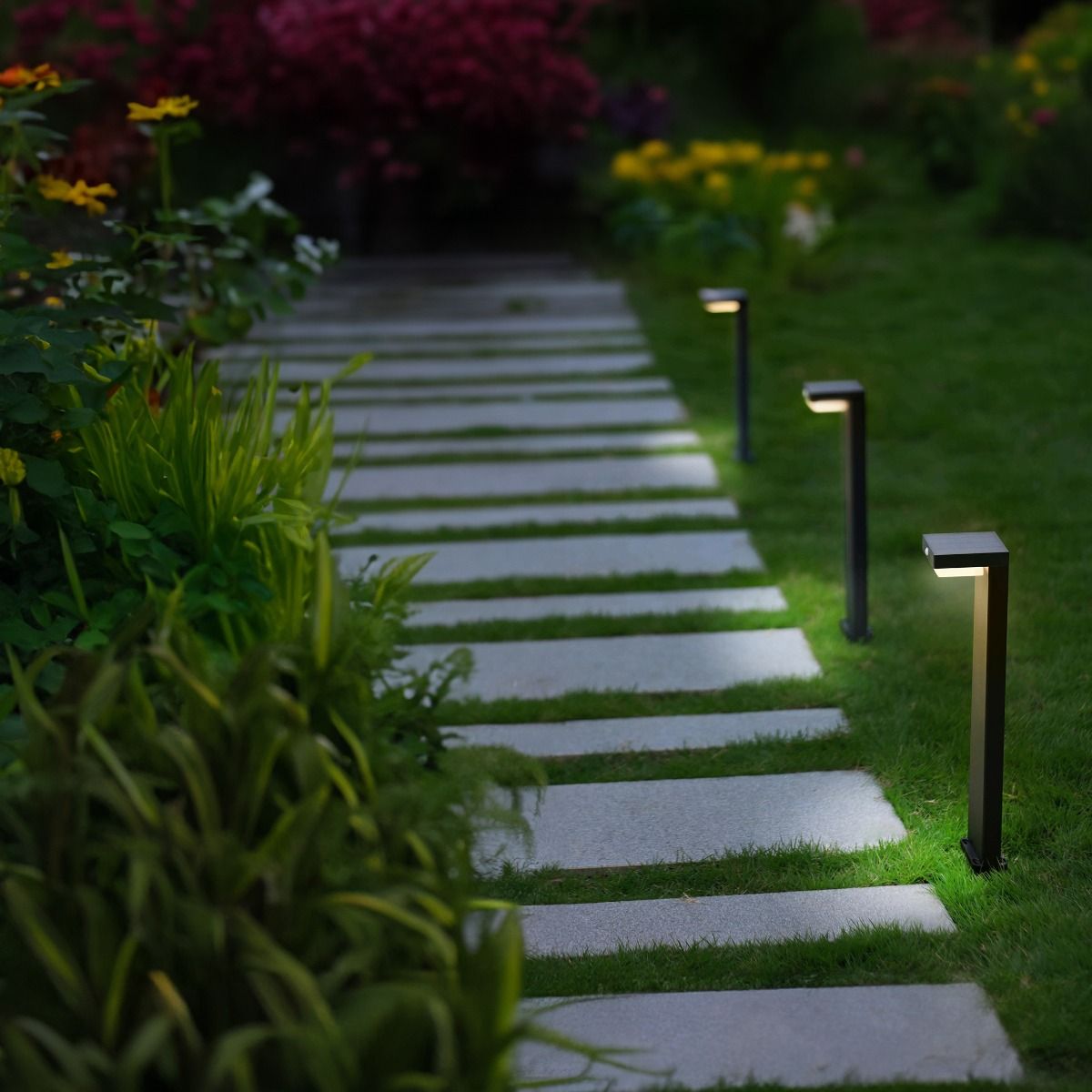 Lampa ogrodowa solarna Grid, czarna, z czujnikiem ruchu, ustawiona na ścieżce z betonowych płytek. Oświetla otoczenie, z widocznymi roślinami w tle, na trawie. Wysoka na około 50 cm, z prostokątną głowicą LED.