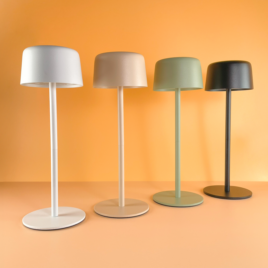 Lampa stołowa LED Poco 2W 3000K IP44 w zielonym kolorze, z okrągłą podstawą i cylindrycznym kloszem, prezentująca minimalistyczny design. Ujęcie frontalne, obok lamp w kolorze białym, beżowym i czarnym, na tle pomarańczowej ściany.