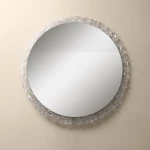 Lustro podświetlane w kształcie koła o średnicy 60 cm, z ramą z kryształowego materiału. W centralnej części gładka powierzchnia lustra, z delikatnym włącznikiem dotykowym u dołu. Zawieszone na jasnym tle.