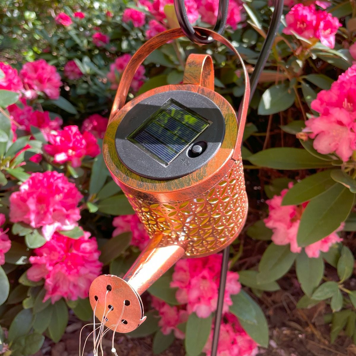Konewka lampa solarna LED o mocy 0,06W, kolor czarny/miedź, z widocznym panelem słonecznym i detalami w formie otworów na końcu wylewki. Zawieszona w ogrodzie, w otoczeniu różowych kwiatów.