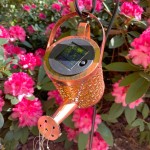 Konewka lampa solarna LED o mocy 0,06W, kolor czarny/miedź, z widocznym panelem słonecznym i detalami w formie otworów na końcu wylewki. Zawieszona w ogrodzie, w otoczeniu różowych kwiatów.