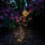 Konewka lampa solarna LED w kolorze czarnym i miedzianym, umieszczona na stojaku, emituje ciepłe światło 3000K. Dookoła widoczne kwiaty różaneczników, a lampa o wzorze perforowanym zdobi otoczenie, tworząc romantyczny klimat.