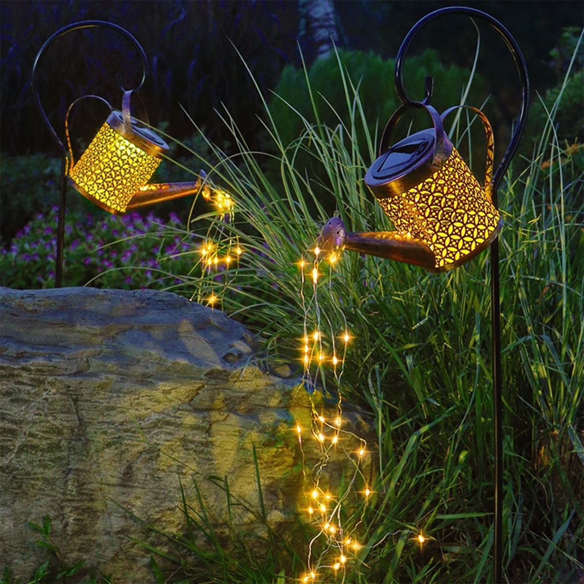 Konewka lampa solarna LED w kolorze czarnym i miedzianym, umieszczona na metalowym stelażu. Emituje ciepłe światło 3000K, ozdobiona wzorami. W tle trawa oraz kamień, a z konewki spływają drobne lampki LED. Idealna do ogrodu.