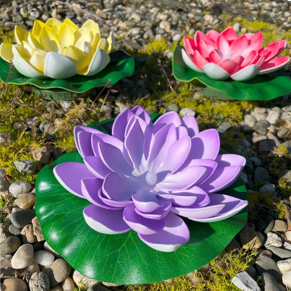 Lampa solarna LED w kształcie kwiatu lotosu w kolorze czerwonym, umieszczona na zielonym liściu. Widoczne detale płatków i środek lampy. Otoczenie z kamieni i mchu, na tle naturalnym. Idealna do ogrodu lub na taras.