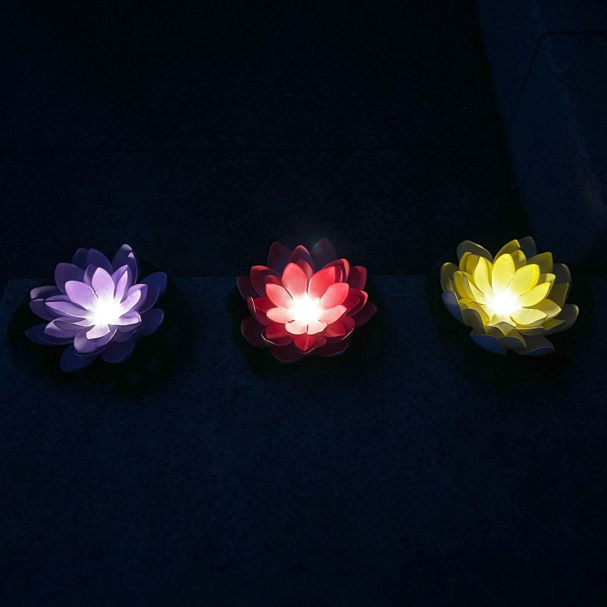 Lampa solarna LED w kształcie kwiatu lotosu, czerwona, o mocy 0,06W i barwie światła 3000K. Na zdjęciu trzy lampy w różnych kolorach: fioletowa, czerwona i żółta, emitujące delikatne światło, umieszczone na ciemnym tle.