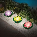 Lampa solarna LED w kształcie kwiatu lotosu, żółta, z zielonym podłożem. Widoczne trzy kwiaty: żółty, różowy i fioletowy, umieszczone na białych kamieniach, otoczone roślinnością. Oświetlenie ciepłe, 3000K, IP44.
