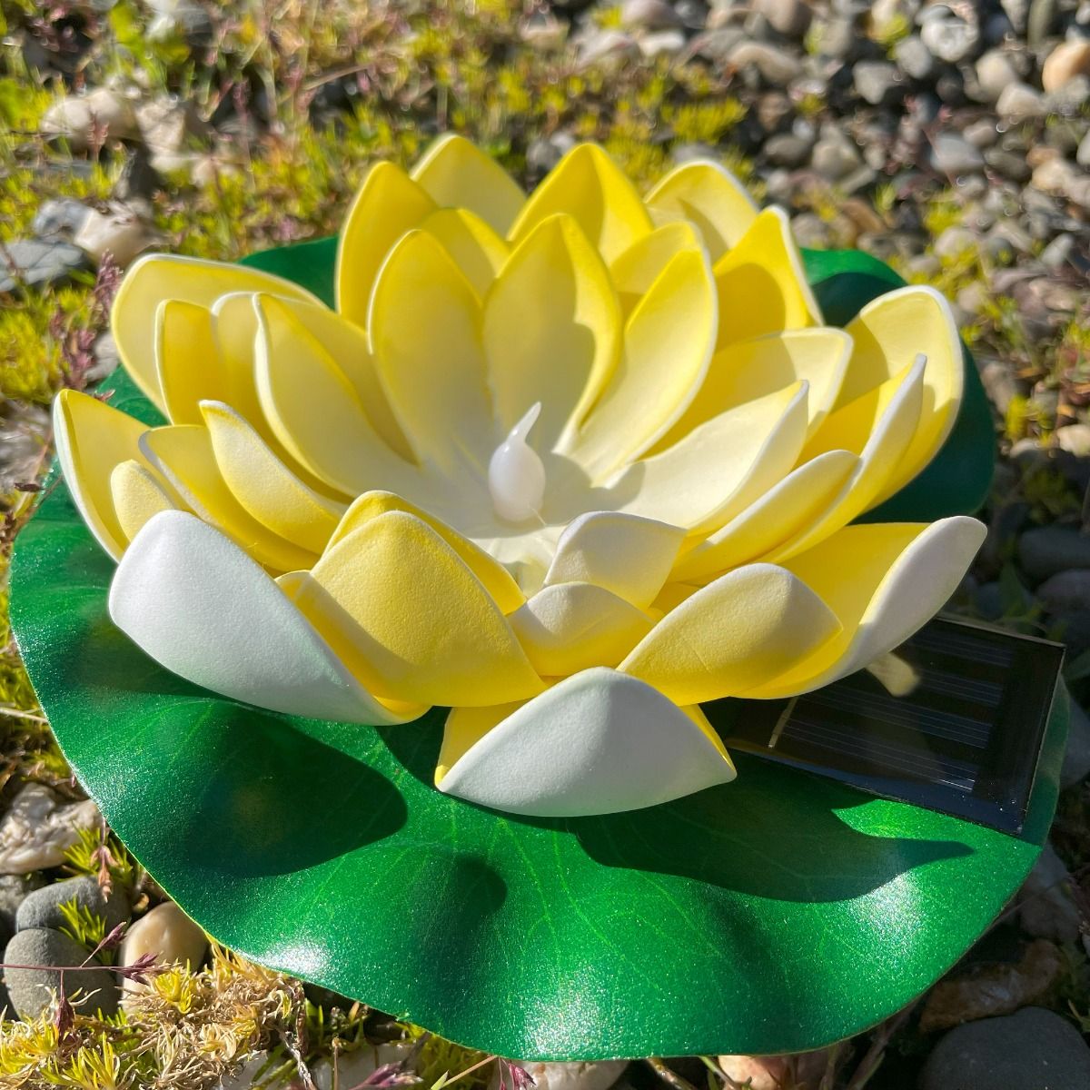 Lampa solarna LED w kształcie kwiatu lotosu, żółta, z białymi detalami. Umieszczona na zielonym liściu, z widocznym panelem solarnym. Produkt leży na ziemi w naturalnym otoczeniu. Wymiary: średnica kwiatu około 20 cm. IP44, moc 0,06W, barwa 3000K.