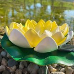 Lampa solarna LED w kształcie kwiatu lotosu, z żółtymi i białymi płatkami, umieszczona na zielonym liściu. Widać elementy montażowe. Produkt leży na kamieniach, w tle odbicie wody. IP44, 0,06W, 3000K.