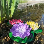 Lampa solarna LED w kształcie fioletowego kwiatu lotosu na zielonym liściu, z widocznymi sokowymi detalami. W tle staw z kamieniami, obok czerwony i żółty kwiat lotosu. Produkt o mocy 0,06W, temperatura barwowa 3000K, stopień ochrony IP44.