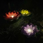 Lampa solarna LED w kształcie kwiatu lotosu, fioletowa, z delikatnym podświetleniem. W otoczeniu zielonych liści i trawy, z widocznymi czerwonym i żółtym kwiatem obok. Ochrona IP44, moc 0,06W, temperatura barwowa 3000K.