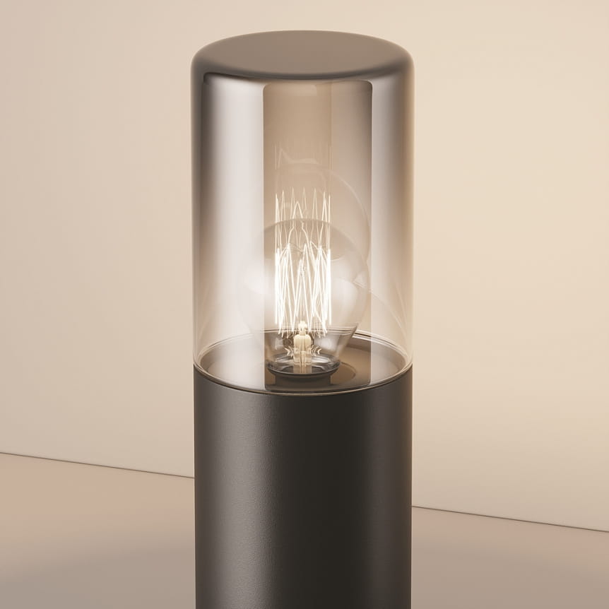 Lampa ogrodowa Amas H25 w kolorze grafitowym, z cylindrycznym korpusem i przezroczystą osłoną. Widoczna żarówka E27 z dekoracyjnym filamentem, stojąca na prostym tle.