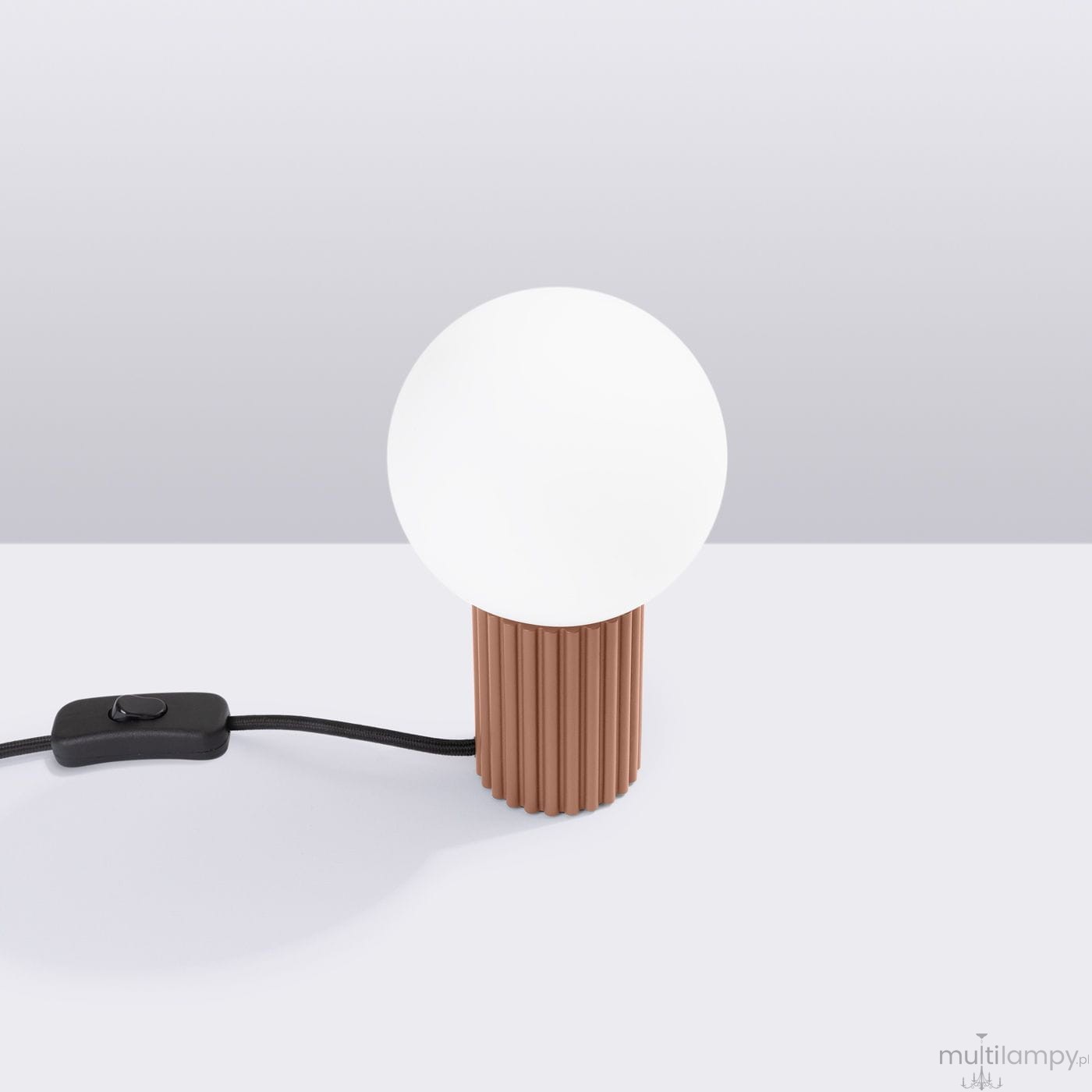Lampa stołowa Halo z okrągłym, białym kloszem i cylindryczną podstawą w kolorze mocca. Widoczny czarny przewód z przełącznikiem. Ujęcie z boku na jasnym tle, podkreślające teksturę podstawy.