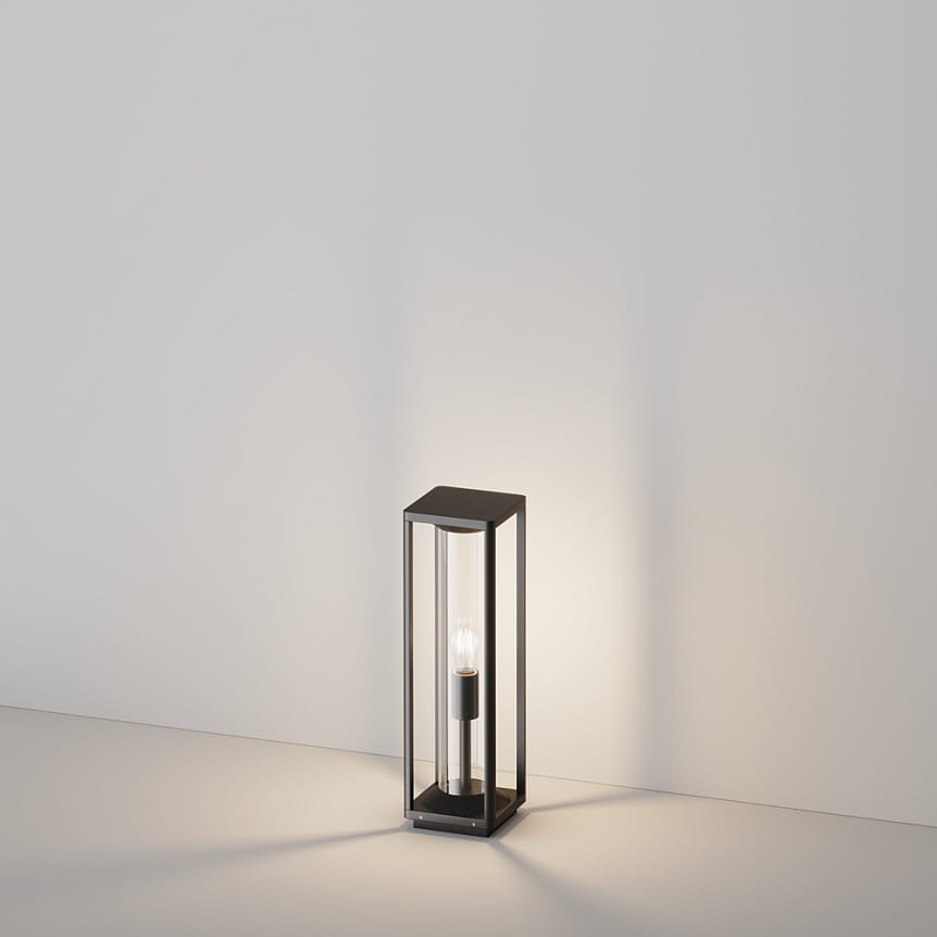Stojąca lampa ogrodowa Cell H50 w grafitowej obudowie, prostokątna konstrukcja z przezroczystym kloszem, umieszczona na płaskiej powierzchni. Widoczne źródło światła E27 w centralnej części, emitujące ciepłe światło.