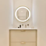 Lustro podświetlane D60 w kształcie koła, z błyszczącą złotą ramą i oświetleniem LED. Zawieszone nad minimalistyczną umywalką z szufladami. Tło jasne, w nowoczesnej łazience. Widoczne akcesoria na blacie, w tym kosmetyki i pojemniki.