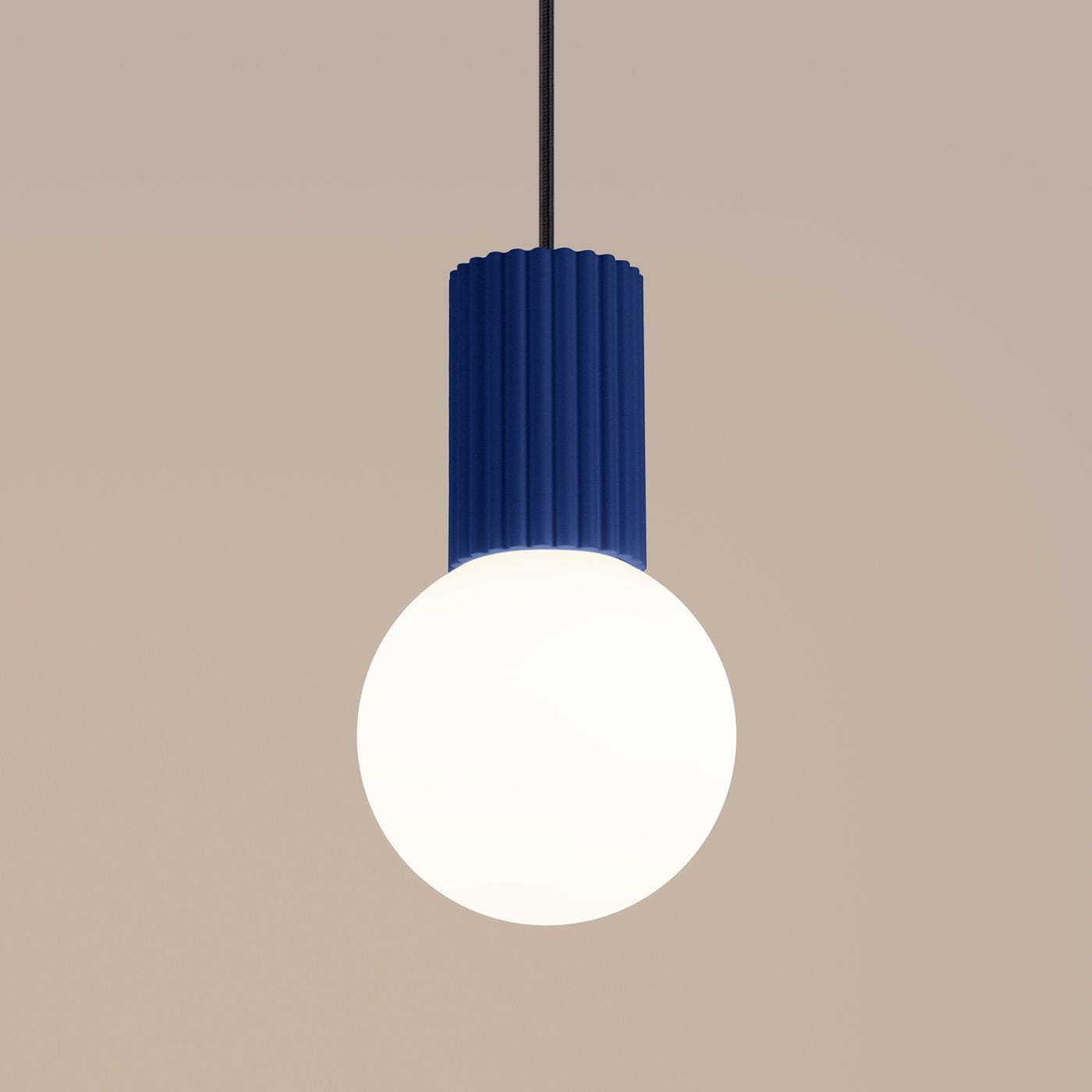 Lampa wisząca Halo z niebieskim, teksturowanym kloszem w formie cylindrycznej oraz białą, okrągłą żarówką. Zawieszona na cienkim, czarnym przewodzie. Tło w jasnym odcieniu.