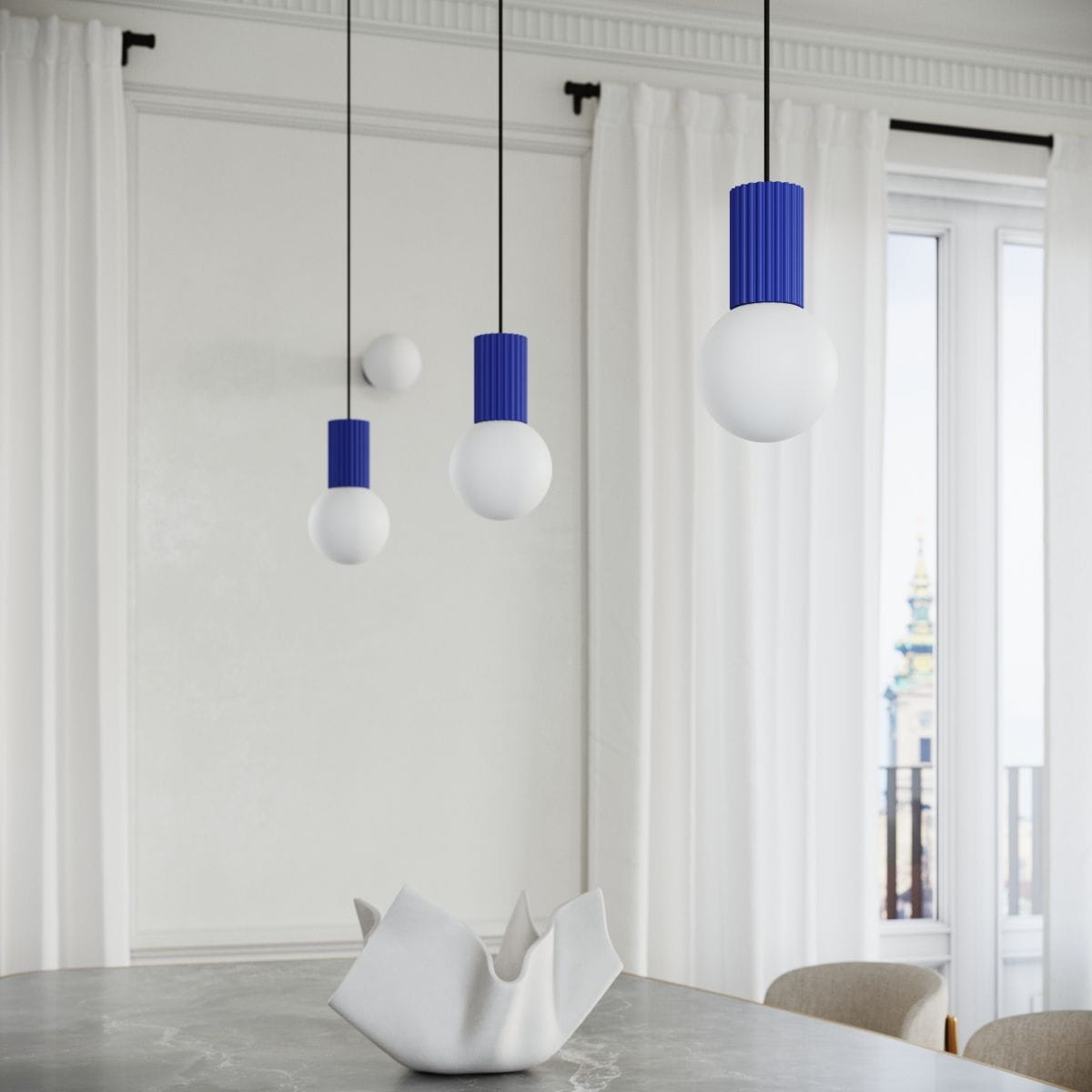 Lampa wisząca Halo z jedną żarówką G9, z okrągłymi kloszami w białym kolorze i niebieskimi, żebrowanymi mocowaniami. Zawieszona na czarnych linkach, w aranżacji na tle jasnych zasłon i okna. Na stole znajduje się ceramiczny element dekoracyjny.