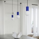 Lampa wisząca Halo z jedną żarówką G9, z okrągłymi kloszami w białym kolorze i niebieskimi, żebrowanymi mocowaniami. Zawieszona na czarnych linkach, w aranżacji na tle jasnych zasłon i okna. Na stole znajduje się ceramiczny element dekoracyjny.