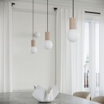 Lampa wisząca Halo w kolorze taupe z jednym źródłem światła G9. Posiada trzy okrągłe klosze z mlecznego szkła, podtrzymywane przez drewniane elementy w kształcie cylindrów. Zawieszona nad stołem na tle jasnych zasłon i dużego okna.