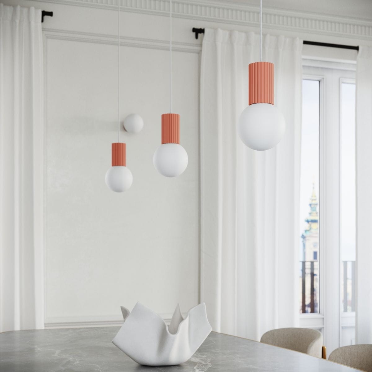 Lampa wisząca Halo w kolorze brzoskwiniowym z białymi kloszami, zawieszona w grupie trzech elementów na cienkich linkach. Widoczny matowy klosz oraz dekoracyjny, prążkowany uchwyt. Lampa znajduje się nad stołem, w jasnym wnętrzu z oknami i zasłonami.