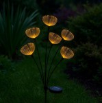 Lampa solarna ogrodowa w kształcie kwiatów, z sześcioma żółtymi, podświetlanymi główkami na cienkich, czarnych łodygach. Ustawiona w pionie, z widocznym panelami słonecznymi na dole, otoczona zieloną trawą i roślinami.