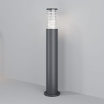 Grafitowa lampa ogrodowa Bronx H80 z jedną oprawą E27. Posiada cylindryczny kształt z przezroczystą osłoną na górze. Ujęcie z boku, produkt stoi na podłożu, z prostą ścianą w tle. Idealna do oświetlenia przestrzeni zewnętrznych.