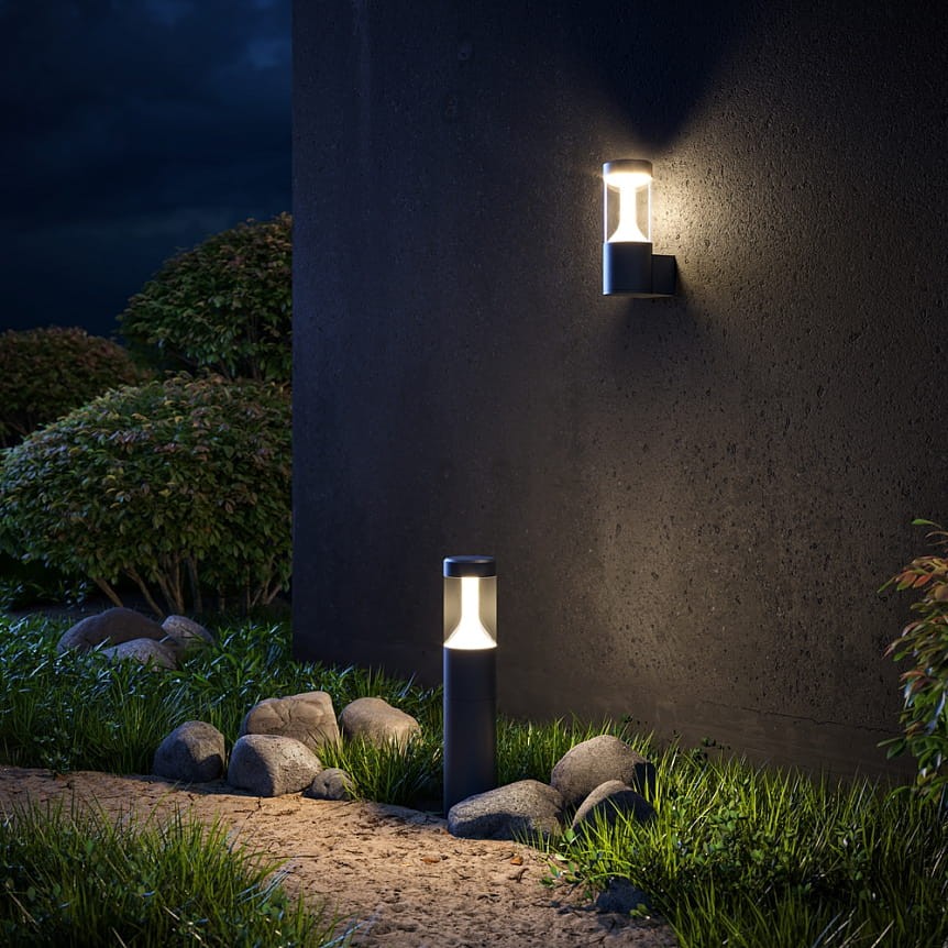 Czarna lampa ogrodowa LED Koln H65 o mocy 8W i temperaturze barwowej 4000K, umieszczona obok ściany. Z przodu widoczna lampa stojąca, a z boku lampa montowana na ścianie, otoczona zieloną trawą i kamieniami.