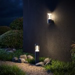 Czarna lampa ogrodowa LED Koln H65 o mocy 8W i temperaturze barwowej 4000K, umieszczona obok ściany. Z przodu widoczna lampa stojąca, a z boku lampa montowana na ścianie, otoczona zieloną trawą i kamieniami.